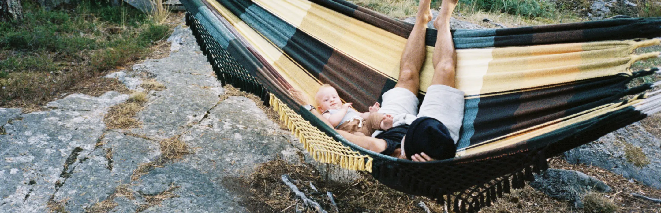 A person and a child lying in a hammock outdoors, surrounded by nature, with a small glasshouse in the background.