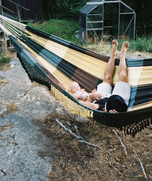 A person and a child lying in a hammock outdoors, surrounded by nature, with a small glasshouse in the background.
